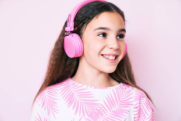 Carino ispanico bambino ragazza ascoltare musica utilizzando cuffie cercando di lato, rilassarsi posa profilo con viso naturale e sorriso fiducioso.  - Foto, immagini