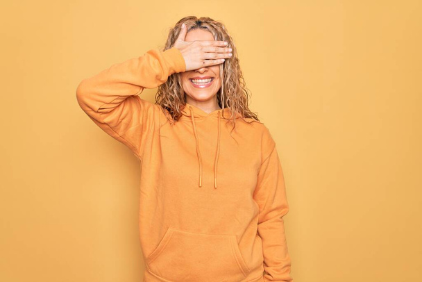 Młoda piękna blondynka sportowa ubrana w luźną bluzę na żółtym tle uśmiechająca się i śmiejąca z ręką na twarzy zakrywająca oczy dla niespodzianki. Koncepcja niewidomych. - Zdjęcie, obraz