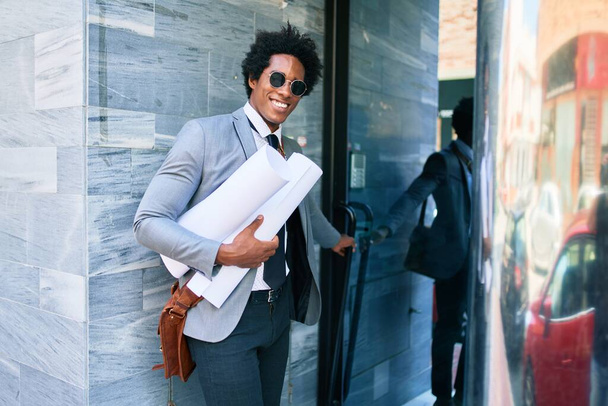 Genç, yakışıklı, Afro-Amerikan mimar mutlu gülümsüyor. Yüzünde bir gülümseme, elinde ofis kapısının planları.. - Fotoğraf, Görsel