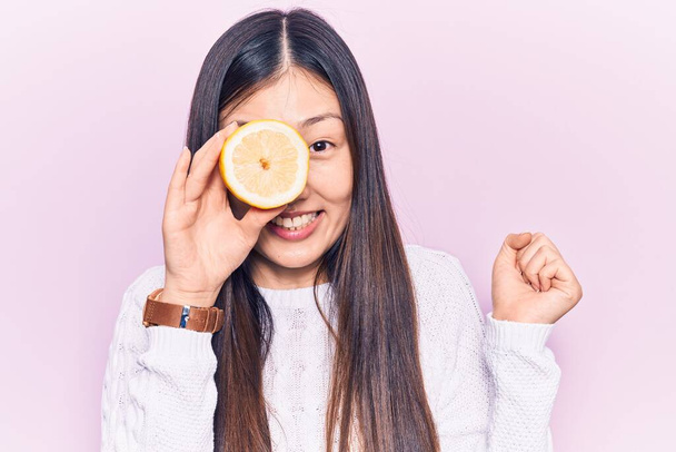 Joven mujer china hermosa sosteniendo rebanada de limón sobre el ojo gritando orgulloso, celebrando la victoria y el éxito muy emocionado con el brazo levantado  - Foto, imagen