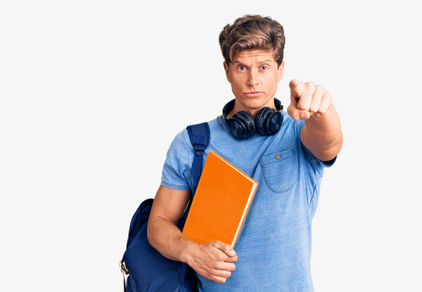 Jeune bel homme portant un sac à dos d'étudiant et un casque tenant un livre pointant du doigt vers la caméra et vers vous, geste confiant et sérieux  - Photo, image