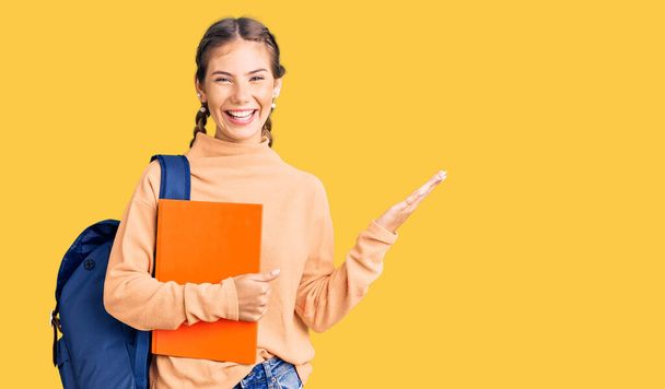 Beautiful caucasian woman with blonde hair wearing student backpack and holding book celebrating victory with happy smile and winner expression with raised hands  - Photo, Image