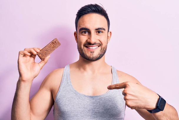 Joven hombre guapo con barba comiendo barra de proteína de energía sobre fondo rosa aislado señalando el dedo a uno mismo sonriendo feliz y orgulloso - Foto, Imagen