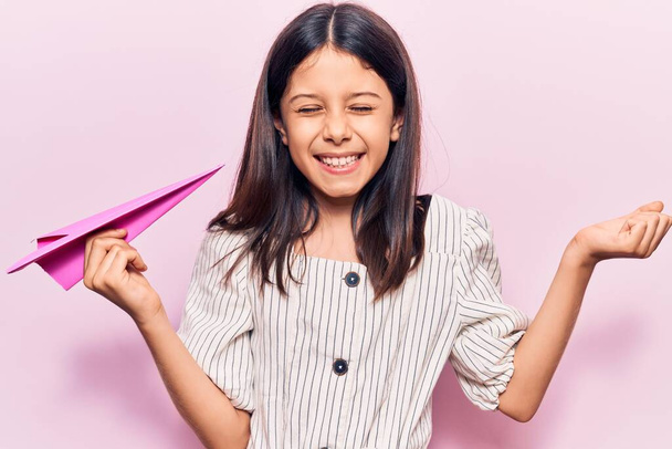 Beautiful child girl holding paper airplane screaming proud, celebrating victory and success very excited with raised arm  - Photo, Image