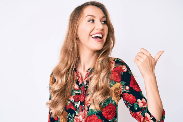 Mulher branca jovem com cabelo loiro vestindo camisa floral elegante apontando polegar até o lado sorrindo feliz com a boca aberta
  - Foto, Imagem