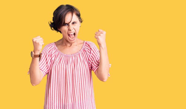 Beautiful young woman with short hair wearing casual summer clothes angry and mad raising fists frustrated and furious while shouting with anger. rage and aggressive concept.  - Photo, image