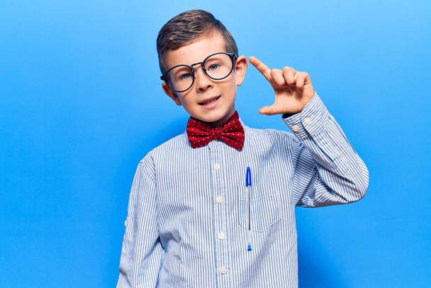 オタクの蝶ネクタイとメガネを身に着けているかわいいブロンドの子供は、指を見て、カメラで小さなサイズの看板を行う手で笑顔と自信を持ってジェスチャー。概念を測定し.  - 写真・画像