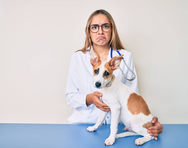 Jonge mooie blonde dierenarts vrouw controleren hond gezondheid sceptisch en nerveus, fronsen boos vanwege probleem. negatieve persoon.  - Foto, afbeelding