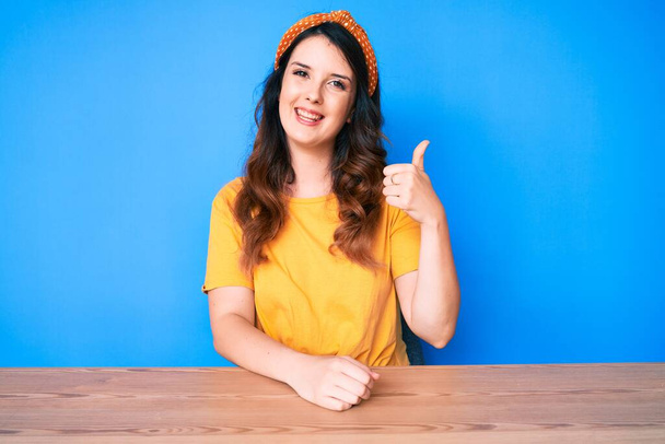 Fiatal gyönyörű barna nő ül az asztalon a kék háttér mosolyogva boldog és pozitív, hüvelykujj fel csinál kiváló és jóváhagyási jel  - Fotó, kép