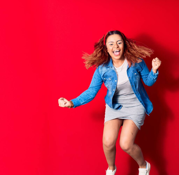 Young beautiful latin woman wearing casual clothes smiling happy. Jumping with smile on face doing winner gesture with fists up over isolated red background - Photo, image