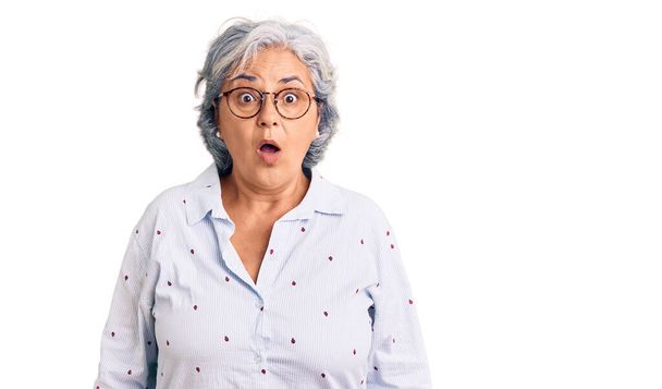 Femme âgée aux cheveux gris portant des vêtements de travail décontractés et des lunettes effrayées et étonnées avec la bouche ouverte pour la surprise, visage incrédule  - Photo, image