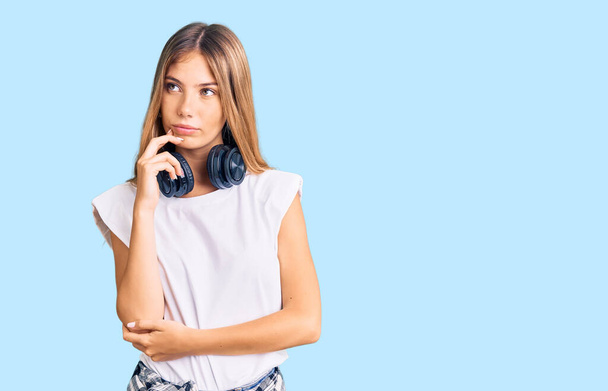 Beautiful caucasian woman with blonde hair listening to music wearing headphones serious face thinking about question with hand on chin, thoughtful about confusing idea  - Foto, afbeelding