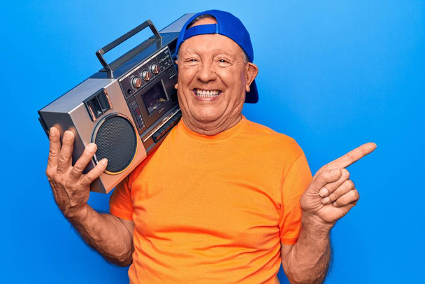 Senior modern jóképű, ősz hajú férfi sapkában zenehallgatás boombox mosoly boldog mutató kéz és ujj az oldalán - Fotó, kép