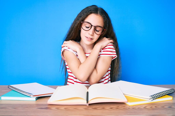 Χαριτωμένο ισπανόφωνο κορίτσι που σπουδάζει για τις σχολικές εξετάσεις κάθεται στο τραπέζι αγκαλιάζοντας τον εαυτό του ευτυχισμένη και θετική, χαμογελώντας αυτοπεποίθηση. αυτοαγάπη και αυτοφροντίδα  - Φωτογραφία, εικόνα