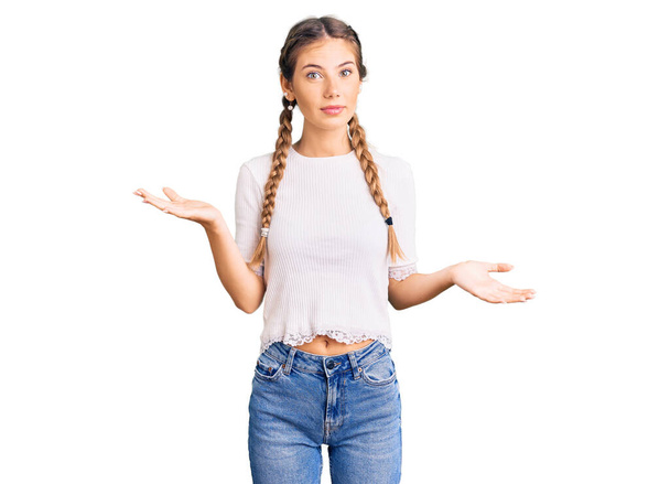 Beautiful caucasian woman with blonde hair wearing braids and white tshirt clueless and confused expression with arms and hands raised. doubt concept.  - Foto, afbeelding