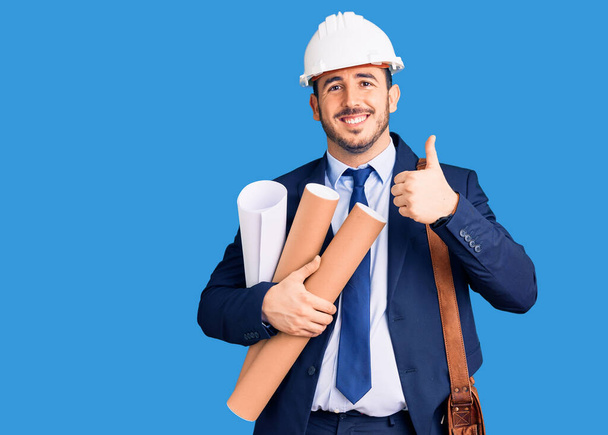 Jonge Spaanse man met een hardhoed van architect en leren tas met blauwdrukken die vrolijk en positief glimlacht, duim omhoog doet het uitstekend en goedkeuringsteken  - Foto, afbeelding