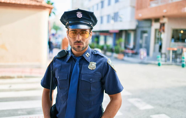 Jeune beau policier hispanique en uniforme de police. Debout avec expression sérieuse dans la rue de la ville. - Photo, image
