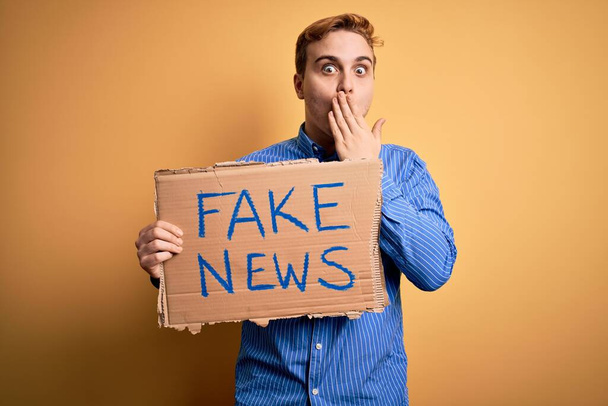 Junge hübsche rothaarige Mann mit Banner mit Fake-News-Nachricht über gelbem Hintergrund bedeckt Mund mit der Hand, schockiert und Angst vor Fehler. Überraschte Miene - Foto, Bild