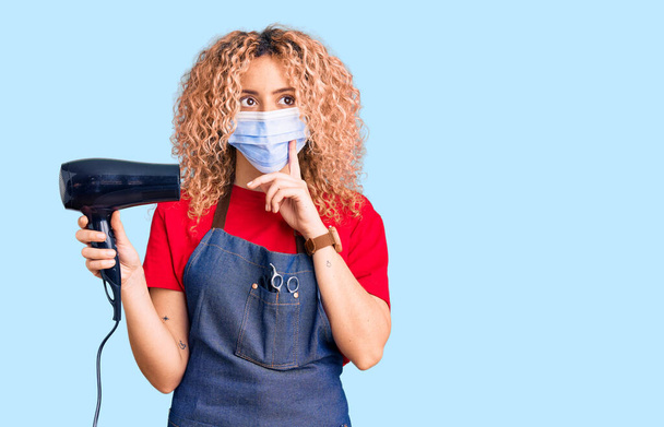 Young blonde woman with curly hair holding dryer blow wearing safety mask for coranvirus serious face thinking about question with hand on chin, thoughtful about confusing idea  - Photo, Image
