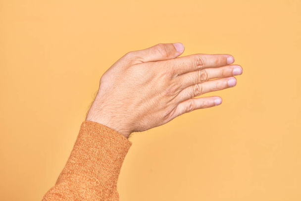 Χέρι καυκάσιου νεαρού άνδρα που δείχνει τα δάχτυλά του πάνω από απομονωμένο κίτρινο φόντο που εκτείνεται και φτάνει με ανοιχτό χέρι για χειραψία, δείχνοντας το πίσω μέρος του χεριού - Φωτογραφία, εικόνα