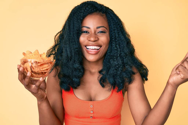 Beautiful african woman holding nachos potato chips screaming proud, celebrating victory and success very excited with raised arms  - Photo, image