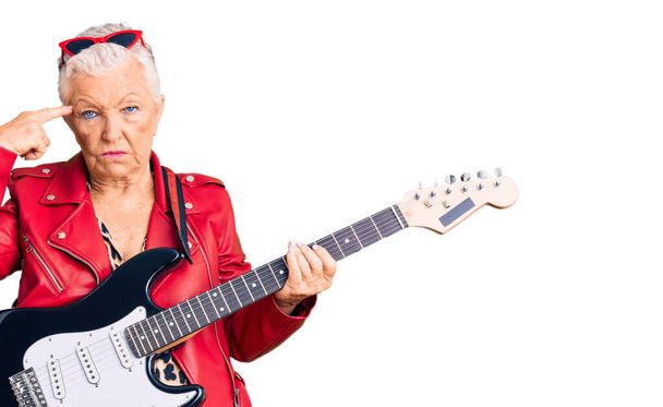 Senior όμορφη γυναίκα με μπλε μάτια και γκρίζα μαλλιά φορώντας ένα μοντέρνο βλέμμα παίζει ηλεκτρική κιθάρα γυρίσματα και σκοτώνοντας τον εαυτό δείχνει το χέρι και τα δάχτυλα στο κεφάλι σαν όπλο, χειρονομία αυτοκτονίας.  - Φωτογραφία, εικόνα
