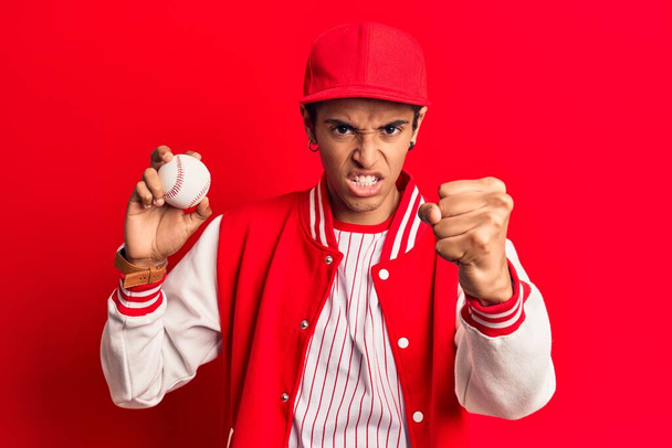 Joven africano amerciano vestido con uniforme de béisbol sosteniendo la pelota molesto y frustrado gritando con ira, gritando loco de ira y la mano levantada  - Foto, imagen