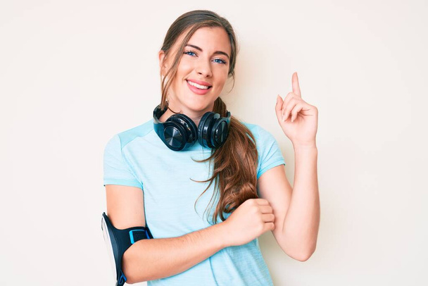 Beautiful young caucasian woman wearing gym clothes and using headphones with a big smile on face, pointing with hand and finger to the side looking at the camera.  - Photo, Image