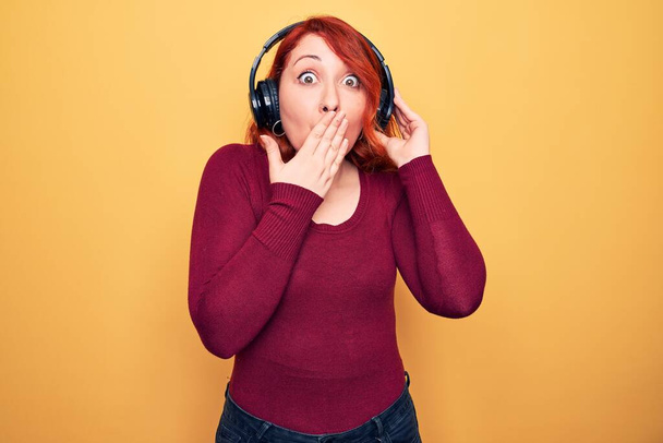Young beautiful redhead woman listening to music using headphones over yellow background covering mouth with hand, shocked and afraid for mistake. Surprised expression - Fotografie, Obrázek