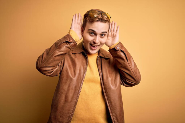 Young handsome redhead man wearing casual leather jacket over isolated yellow background Smiling cheerful playing peek a boo with hands showing face. Surprised and exited - Photo, Image