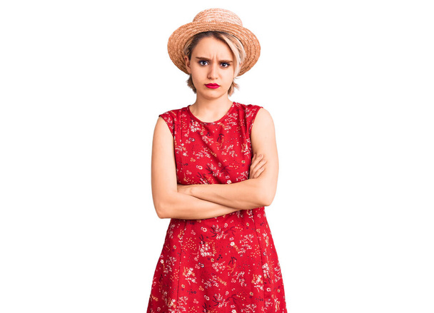 若い美しいブロンドの女性は、夏の帽子懐疑的で緊張し、両腕を組んで顔に不承認の表現を身に着けています。否定的な人.  - 写真・画像