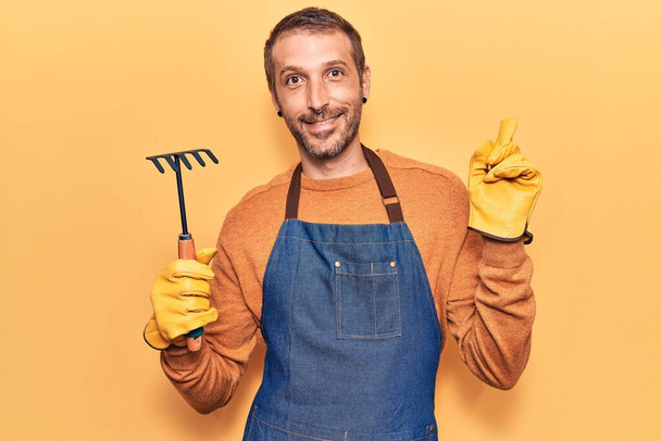 Jonge knappe man draagt tuinschort en handschoenen met hark glimlachend vrolijk wijzend met hand en vinger naar de zijkant  - Foto, afbeelding