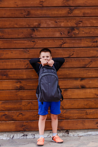 Seven-year-old schoolboy in sportswear holds a heavy backpack outdoors - Фото, изображение