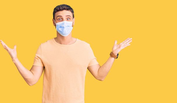 Handsome young man with bear wearing medical mask for coronavirus celebrating victory with happy smile and winner expression with raised hands  - 写真・画像