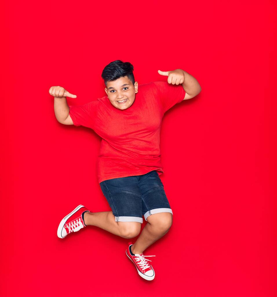 Αξιολάτρευτο παιδί που φοράει καθημερινά ρούχα χαμογελώντας χαρούμενο. Άλμα με χαμόγελο στο πρόσωπο κάνει εντάξει σημάδι με τους αντίχειρες πάνω από απομονωμένο κόκκινο φόντο - Φωτογραφία, εικόνα