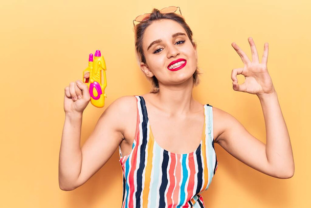Young beautiful blonde woman wearing swimwear holding water gun doing ok sign with fingers, smiling friendly gesturing excellent symbol  - Photo, Image