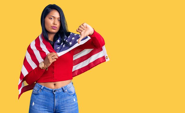 Hispanic woman with long hair holding united states flag with angry face, negative sign showing dislike with thumbs down, rejection concept  - Foto, imagen