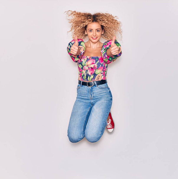 Young beautiful curly girl wearing casual clothes smiling happy. Jumping with smile on face doing ok sign with thumbs up over isolated white background. - 写真・画像