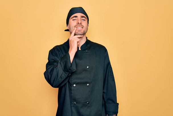 Young handsome cooker man with blue eyes wearing uniform and hat over yellow background looking confident at the camera with smile with crossed arms and hand raised on chin. Thinking positive. - Photo, Image