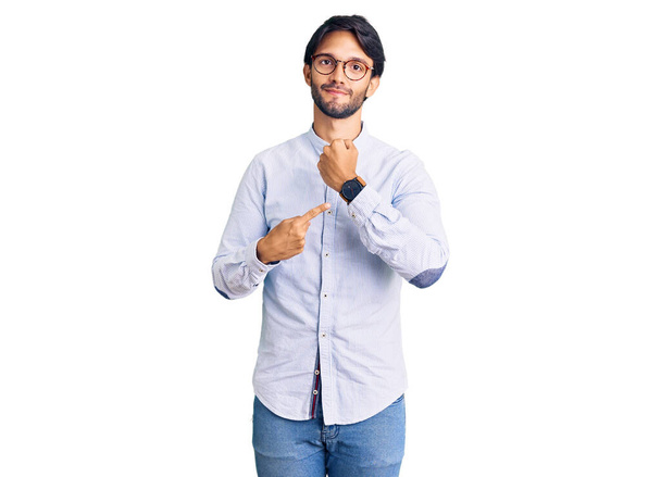 Handsome hispanic man wearing business shirt and glasses in hurry pointing to watch time, impatience, looking at the camera with relaxed expression  - 写真・画像