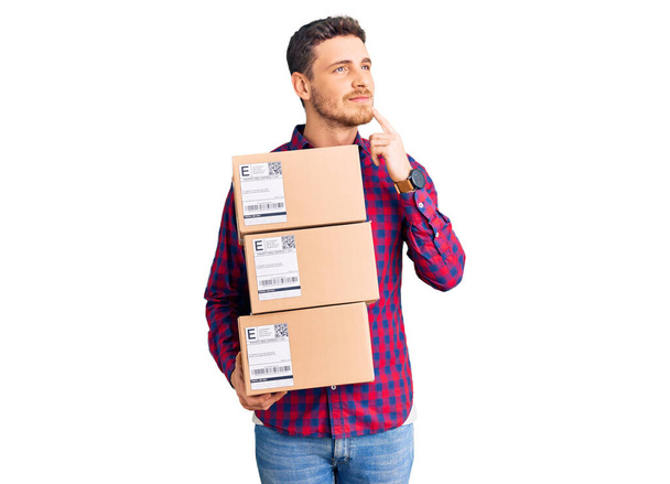 Handsome young man with bear holding delivery packages serious face thinking about question with hand on chin, thoughtful about confusing idea  - Photo, Image