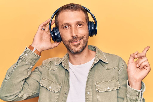 Fiatal jóképű férfi zenét hallgat fejhallgatóval mosolyogva boldog mutogatás kézzel és ujjal oldalra  - Fotó, kép
