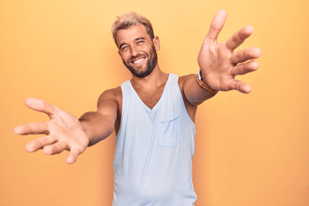 Young handsome blond man wearing casual sleeveless t-shirt over isolated yellow background looking at the camera smiling with open arms for hug. Cheerful expression embracing happiness. - Photo, Image