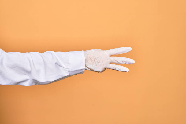 Hand of a person wearing medical surgical glove showing three fingers over isolated yellow background - Photo, image