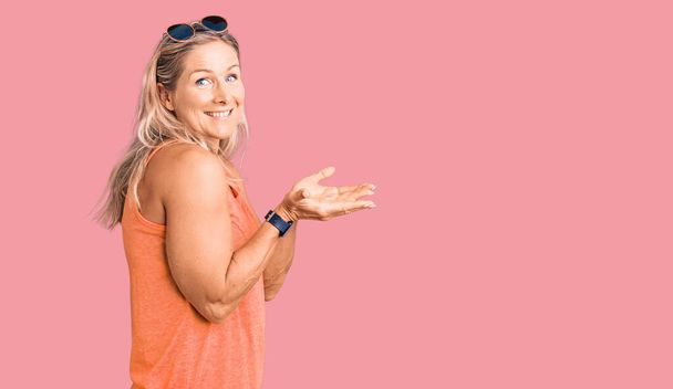 Středního věku fit blondýny žena na sobě ležérní letní oblečení a sluneční brýle ukazuje stranou s rukama otevřené dlaně ukazující kopírovací prostor, prezentující reklamu s nadšeným úsměvem  - Fotografie, Obrázek
