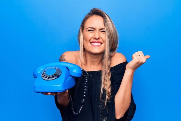 Молода красива блондинка тримає старовинний телефон над ізольованим синім тлом кричить гордо, святкуючи перемогу і успіх дуже схвильований піднятою рукою
 - Фото, зображення