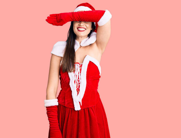 Young beautiful caucasian woman wearing santa claus costume covering eyes with arm smiling cheerful and funny. blind concept.  - Photo, Image