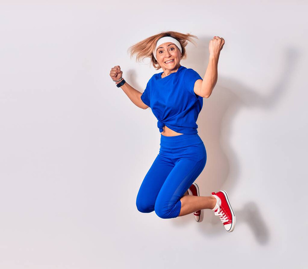 Young beautiful caucasian sportswoman wearing sportswear smiling happy. Jumping with smile on face doing winner sign with fists up over isolated white background - Photo, Image
