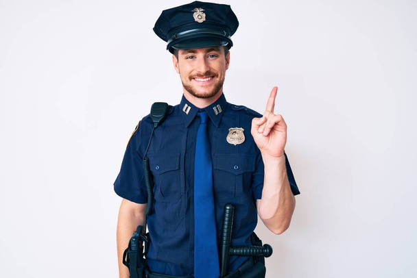 Il giovane caucasico con l'uniforme della polizia punta il dito verso l'alto con un'idea di successo. Uscito e felice. numero uno.  - Foto, immagini
