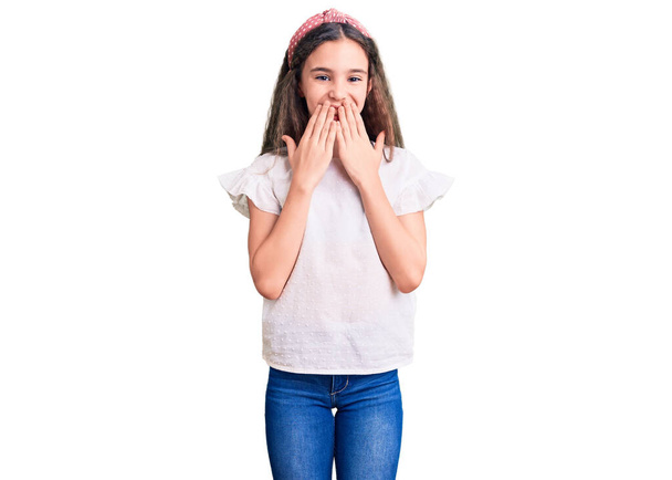 Nettes hispanisches Kindermädchen in lässigem weißem T-Shirt, lachend und verlegen kichernd, den Mund mit Händen bedeckt, Klatsch und Skandalkonzept  - Foto, Bild